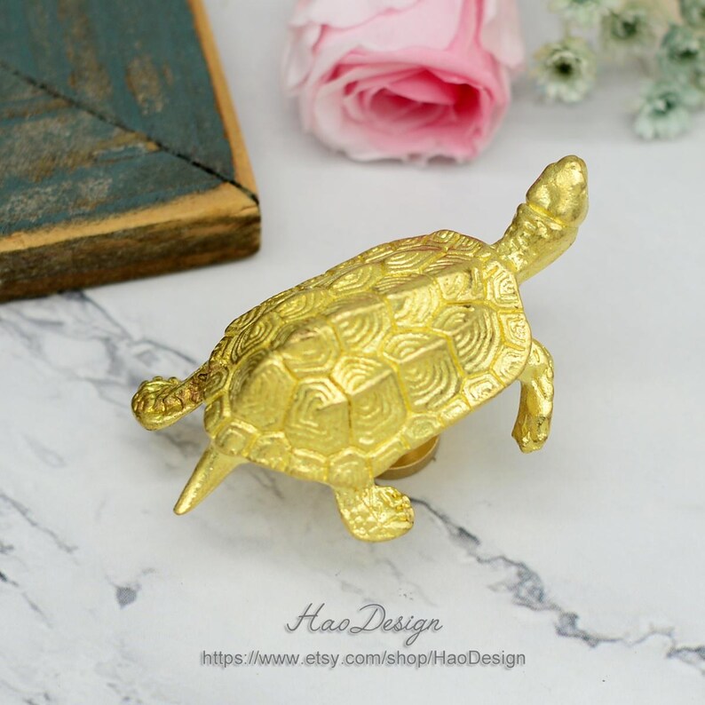 Peut inclure: Une figurine de tortue dor&eacute;e avec un motif de carapace d&eacute;taill&eacute;. La tortue est pos&eacute;e sur une surface en marbre blanc.