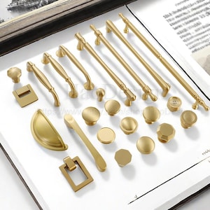 May include: A collection of gold-toned cabinet hardware, including knobs, pulls, and handles, arranged on a white surface. The hardware features various designs, from simple round knobs to ornate pulls, showcasing a range of styles for home decor.