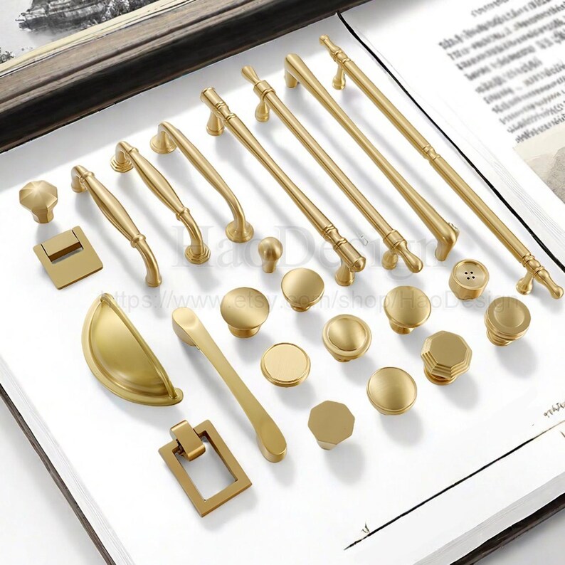 May include: A collection of gold-toned cabinet hardware, including knobs, pulls, and handles, arranged on a white surface. The hardware features various designs, from simple round knobs to ornate pulls, showcasing a range of styles for home decor.
