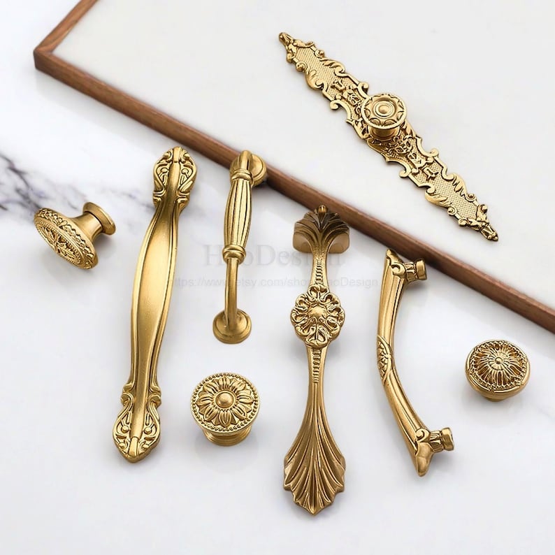 K&ouml;nnte beinhalten: Eine Auswahl an goldenen Schrankbeschl&auml;gen, darunter Kn&ouml;pfe und Griffe. Die Beschl&auml;ge weisen detaillierte Blumen- und Rankenmuster auf. Die Teile variieren in Gr&ouml;&szlig;e und Form und bieten Optionen f&uuml;r verschiedene Schrankstile. Die Ma&szlig;e sind in Zentimetern.