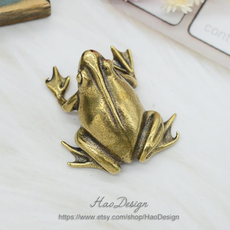 Może przedstawiać: Mała, złota figurka żaby wykonana z mosiądzu. Żaba siedzi z rozłożonymi nogami i uniesioną głową.