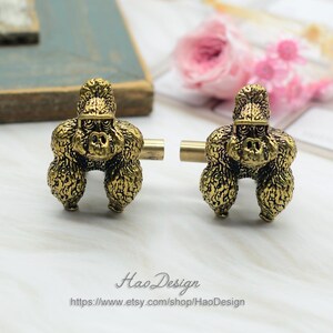 May include: A pair of gold-toned cufflinks featuring a detailed gorilla design. The cufflinks are made of metal and have a textured finish.