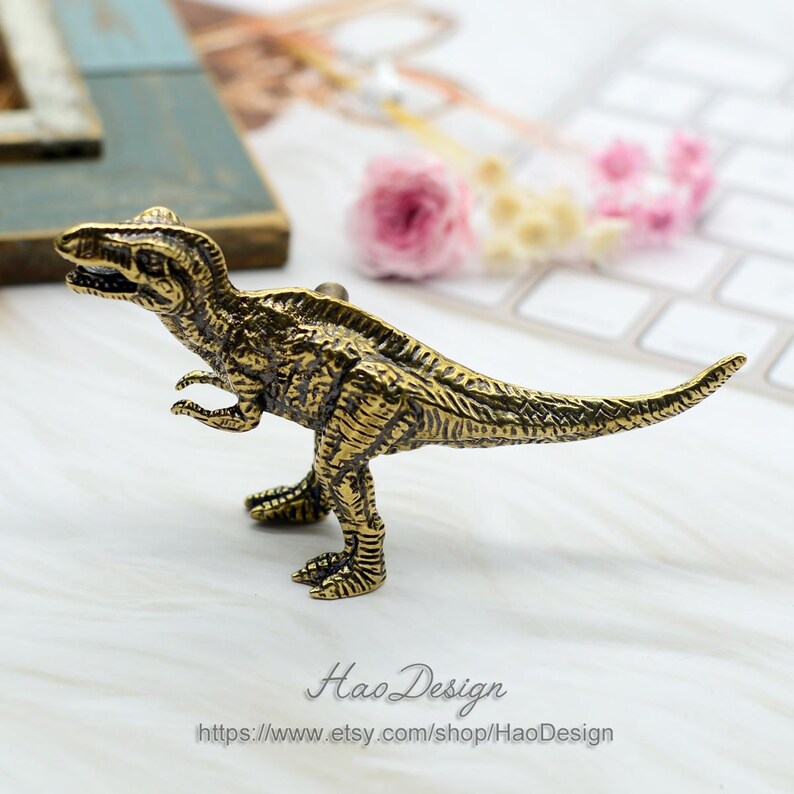 Pode incluir: Uma figura de dinossauro de metal dourado. O dinossauro est&aacute; em p&eacute; sobre uma superf&iacute;cie branca. O dinossauro tem uma superf&iacute;cie texturizada detalhada e uma cauda longa.