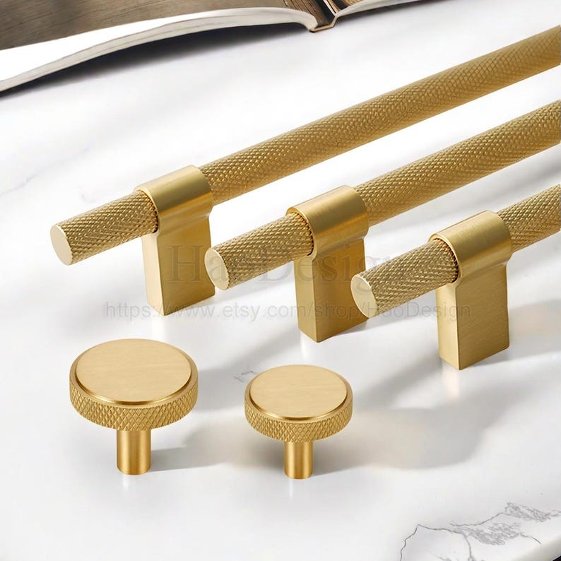 May include: A collection of gold-toned cabinet hardware, including bar pulls and round knobs. The pulls have a textured, diamond-patterned grip and rectangular bases. The knobs have a flat, circular top with a textured edge.