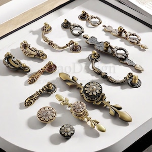 Op de afbeelding: Een assortiment antieke messing kastbeslagen, waaronder handgrepen en knoppen. De hardware heeft sierlijke bloemen- en krulontwerpen, met een warme, verouderde afwerking. Verschillende maten en stijlen worden weergegeven op een wit oppervlak.