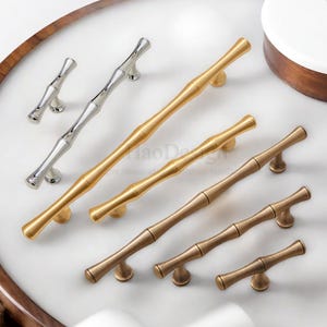 May include: A collection of bamboo-style cabinet pulls in various metallic finishes, including silver, gold, and bronze. The pulls are arranged on a white surface, showcasing their different sizes and designs. The hardware is designed for furniture and cabinetry.