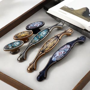 以下が含まれることがあります： 装飾的なキャビネットハードウェアのコレクション。取っ手とノブが含まれています。取っ手は、装飾的なディテールとカラフルな模様のインサートを備えた曲線的なデザインです。ノブも同様のデザインです。ハードウェアはさまざまなメタリック仕上げです。