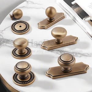 May include: A selection of antique brass cabinet hardware, including knobs and pulls. The knobs have a round design, some with rope detailing. The pulls are rectangular with a rounded top. The hardware is arranged on a white marble surface.