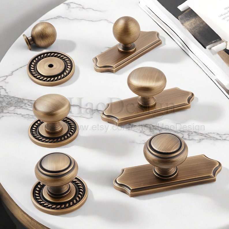 May include: An assortment of antique brass cabinet hardware, including knobs and pulls. The knobs have a round design, some with decorative rope-like detailing. The pulls are rectangular with a rounded top. The hardware is arranged on a white marble surface.