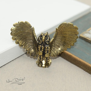 Può includere: Una statuetta di gufo in metallo dorato con le ali spiegate. Il gufo ha un design dettagliato con piume e accenti a forma di perline.