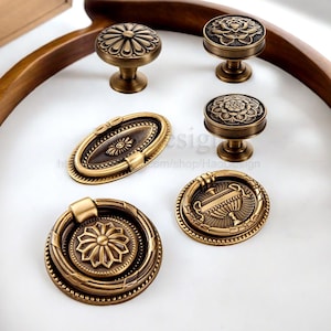 May include: An assortment of antique brass cabinet hardware, including knobs and pulls. The knobs feature floral and geometric designs, while the pulls have oval and ring shapes. The hardware is displayed on a white surface.