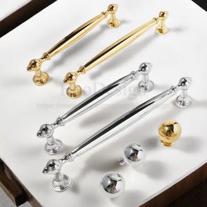 K&ouml;nnte beinhalten: Eine Auswahl an gold- und silberfarbenen Schrankbeschl&auml;gen. Das Bild zeigt zwei goldfarbene Griffe, zwei silberfarbene Griffe und drei Kn&ouml;pfe. Die Griffe haben ein schlankes, l&auml;ngliches Design mit dekorativen Enden. Die Kn&ouml;pfe sind kugelf&ouml;rmig und haben eine polierte Oberfl&auml;che.