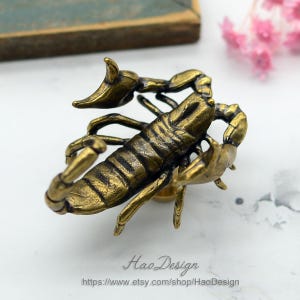 Pode incluir: Uma figurinha de escorpião de metal dourado com textura detalhada. O escorpião está em posição com a cauda levantada e as garras estendidas. É uma peça decorativa que pode ser usada como puxador de gaveta ou como um acento decorativo.