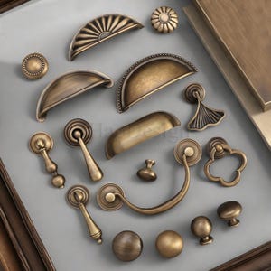 Peut inclure: Une collection de quincaillerie d'armoire en laiton antique, comprenant des boutons, des poignées et des tirettes. La quincaillerie présente divers motifs, tels que des formes en éventail, florales et arrondies. La quincaillerie est disposée sur une surface grise.