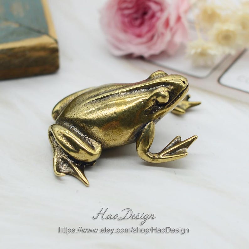 Może przedstawiać: Mała, złota figurka żaby. Żaba siedzi na białym tle. Figurka jest szczeg&oacute;łowa i realistyczna.