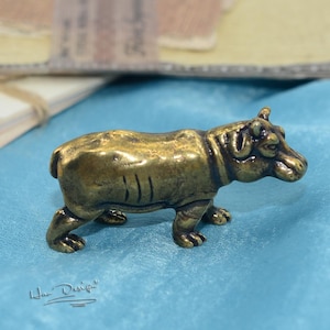 May include: A small, gold-colored metal hippopotamus figurine. The figurine is standing on a blue surface.