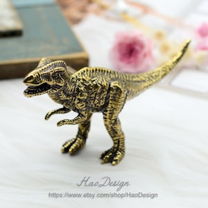 May include: A gold-colored metal dinosaur figurine. The dinosaur is standing on a white surface. The dinosaur is a Tyrannosaurus Rex.