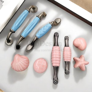 May include: A selection of cabinet hardware. Three light blue ceramic handles with antique bronze details, alongside pink ceramic knobs and pulls in seashell, round, and starfish designs.