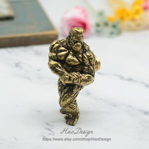May include: A gold-coloured metal cabinet knob shaped like a muscular gorilla. The gorilla is standing with its arms crossed and has a detailed, textured surface.