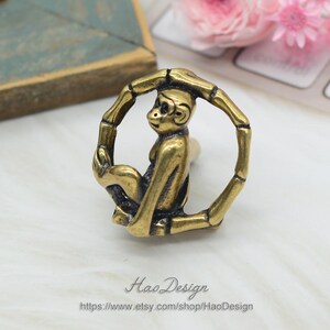 Puede incluir: Una figurita de mono de color dorado sentada dentro de un marco circular de bamb&uacute;. El mono se agarra al marco con sus manos y pies. El marco est&aacute; hecho de finas piezas redondas de bamb&uacute;.