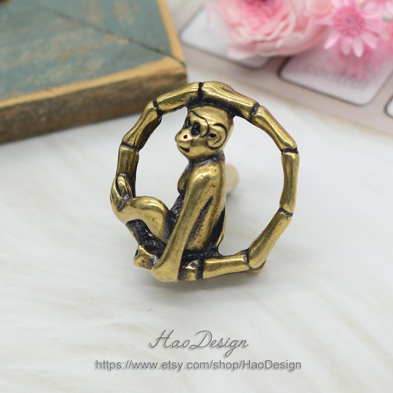 Puede incluir: Una figurita de mono de color dorado sentada dentro de un marco circular de bamb&uacute;. El mono se agarra al marco con sus manos y pies. El marco est&aacute; hecho de finas piezas redondas de bamb&uacute;.