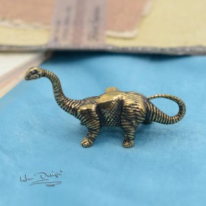 以下が含まれることがあります： 長い首と尾を持つ、小さく詳細な真鍮製の恐竜の置物。 恐竜は4本の足で立っており、質感のある肌と小さな頭があります。 フィギュアの長さは約5cmです。