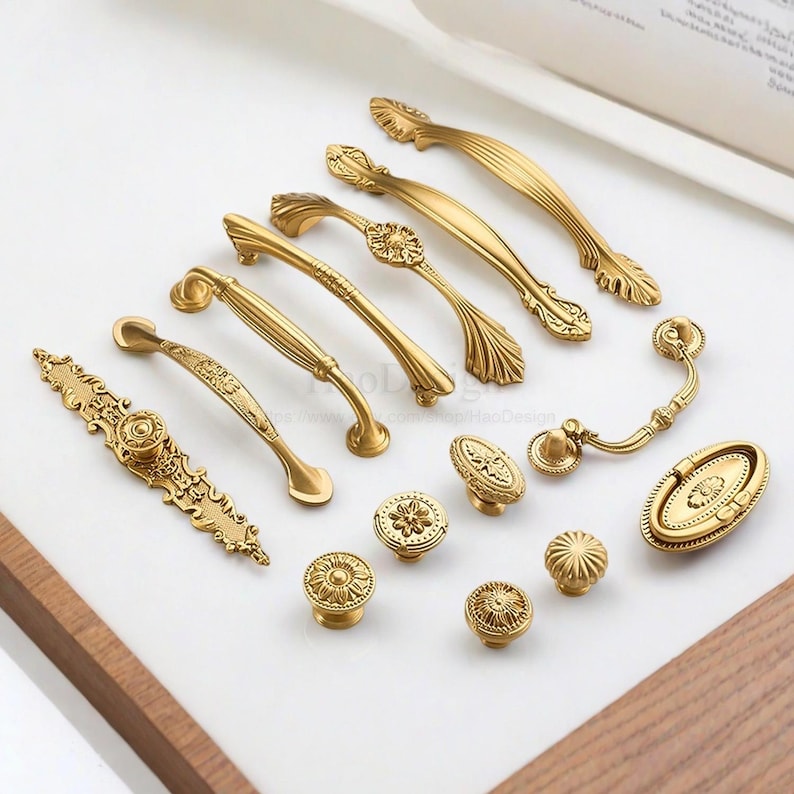 K&ouml;nnte beinhalten: Eine Auswahl an goldenen Schrankbeschl&auml;gen, darunter Griffe und Kn&ouml;pfe. Die Beschl&auml;ge weisen detaillierte florale und geometrische Designs auf und sind in verschiedenen Formen und Gr&ouml;&szlig;en auf einer wei&szlig;en Oberfl&auml;che angeordnet.