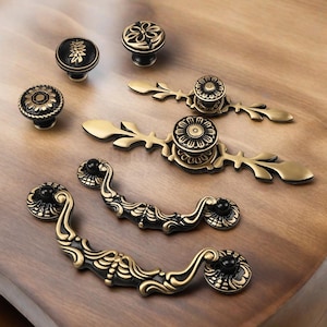 Op de afbeelding: Een assortiment antieke kastbeslagen in zwart en goud. De collectie omvat decoratieve knoppen en handgrepen met bloemen- en bladerpatronen. De beslagen zijn gerangschikt op een houten oppervlak, waarbij de ingewikkelde details en de vintage esthetiek worden benadrukt.