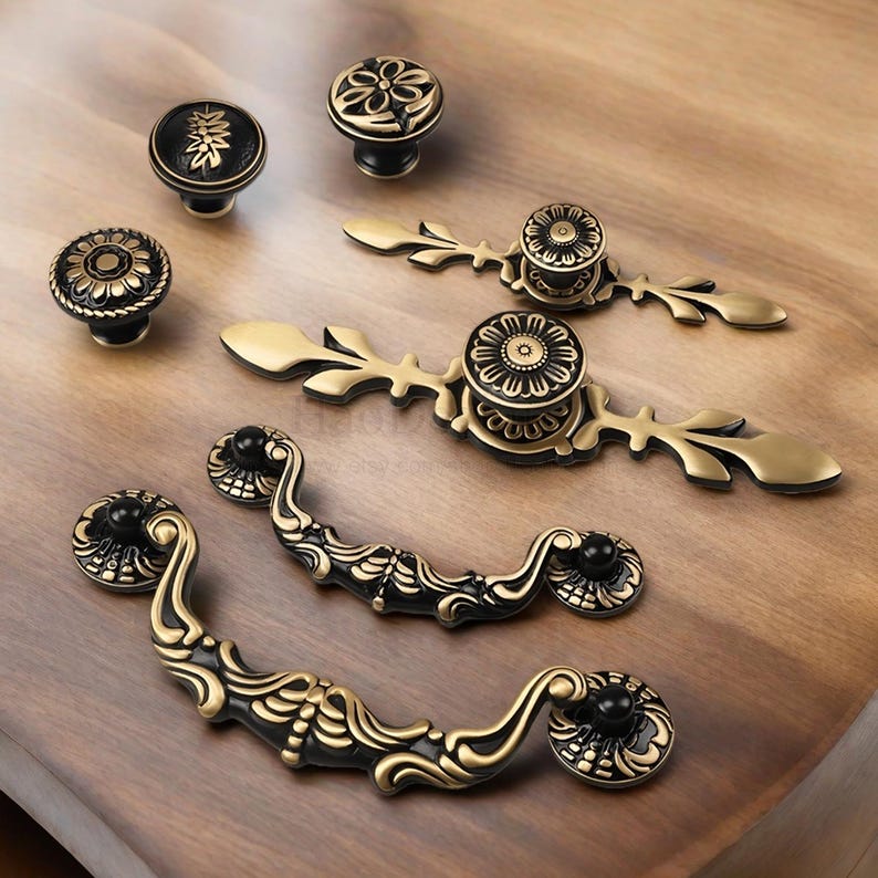 Op de afbeelding: Een assortiment antieke kastbeslagen in zwart en goud. De collectie omvat decoratieve knoppen en handgrepen met bloemen- en bladerpatronen. De beslagen zijn gerangschikt op een houten oppervlak, waarbij de ingewikkelde details en de vintage esthetiek worden benadrukt.