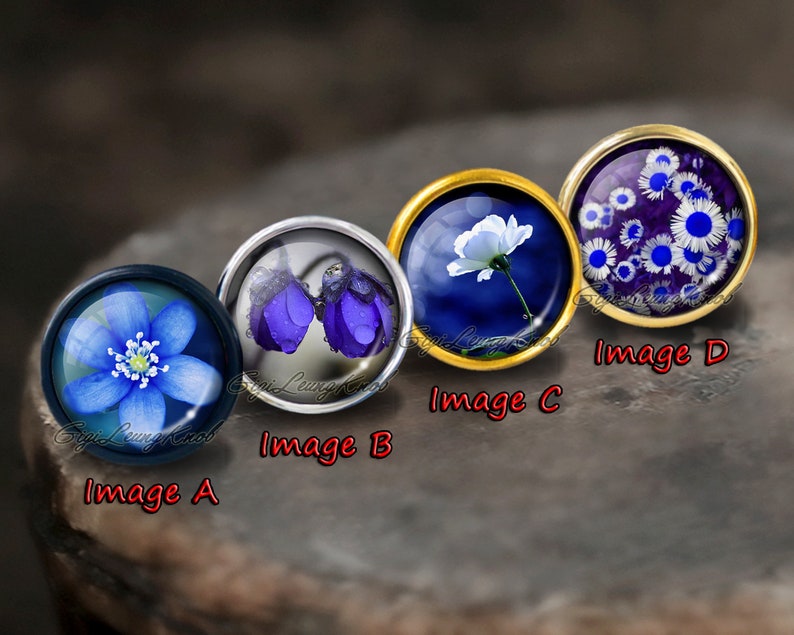 K&ouml;nnte beinhalten: Vier runde Glascabochons mit verschiedenen Blumendesigns. Die Cabochons sind in Metallfassungen gefasst und auf einer dunkelgrauen Oberfl&auml;che ausgestellt. Die Cabochons zeigen eine blaue Blume, zwei lila Blumen, eine wei&szlig;e Blume und eine Ansammlung wei&szlig;er Blumen auf einem lila Hintergrund.