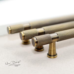 Puede incluir: Tres tiradores de armario de latón antiguo con un patrón de diamante texturizado. Los tiradores tienen un pomo redondo y una barra recta.