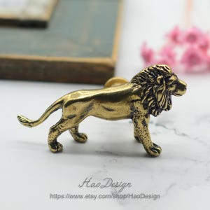 May include: A gold lion figurine door knob or drawer pull. The lion is standing on all four legs with its head turned to the right. The lion has a mane and a tail.