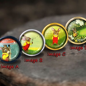 May include: Four round glass cabochons with different images of a rabbit in various settings. The cabochons are set in metal settings with a gold, silver, and black finish. The images include a rabbit under an umbrella, a rabbit holding a dandelion, a rabbit standing in a field, and a rabbit in a field of flowers.