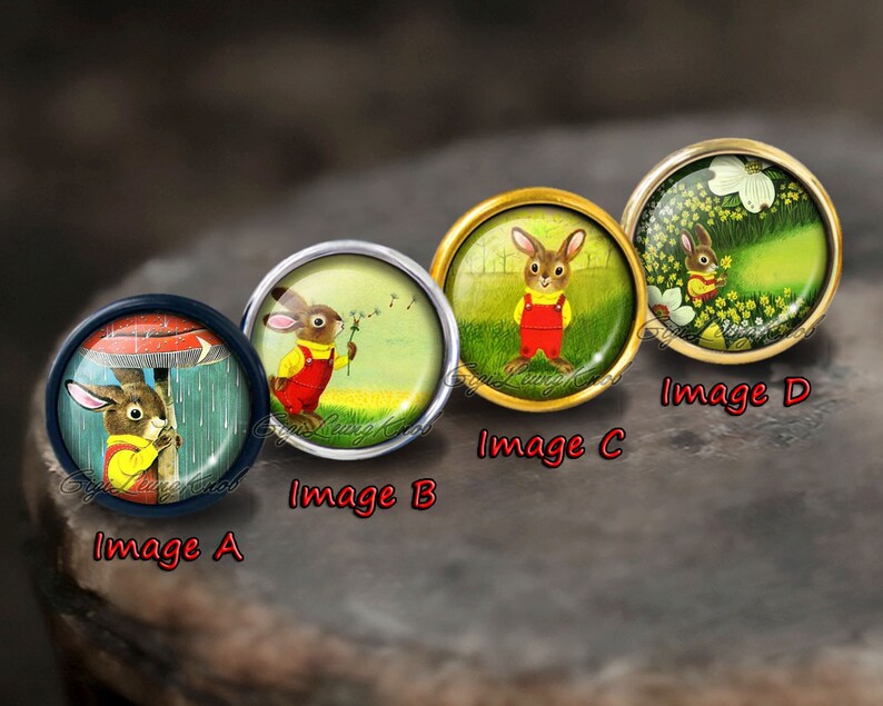 May include: Four round glass cabochons with different images of a rabbit in various settings. The cabochons are set in metal settings with a gold, silver, and black finish. The images include a rabbit under an umbrella, a rabbit holding a dandelion, a rabbit standing in a field, and a rabbit in a field of flowers.