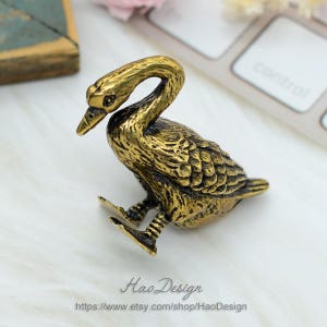 May include: A small, gold-colored metal figurine of a goose with its head turned to the side. The goose is sitting on its feet with its wings folded. The figurine is detailed with textured feathers.