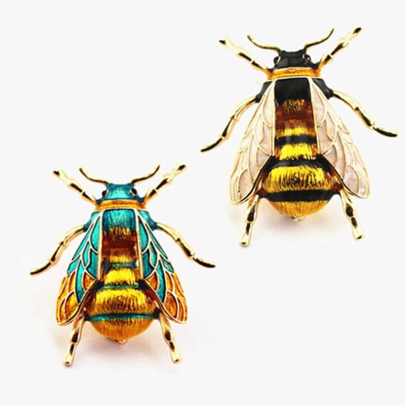 Op de afbeelding: Twee gouden bijen broche met zwarte en gele strepen. E&eacute;n bij heeft turquoise vleugels en de andere heeft witte vleugels.