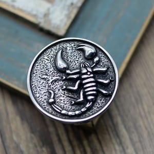 May include: A silver-toned metal ring featuring a detailed scorpion design. The scorpion is raised and has a textured surface.