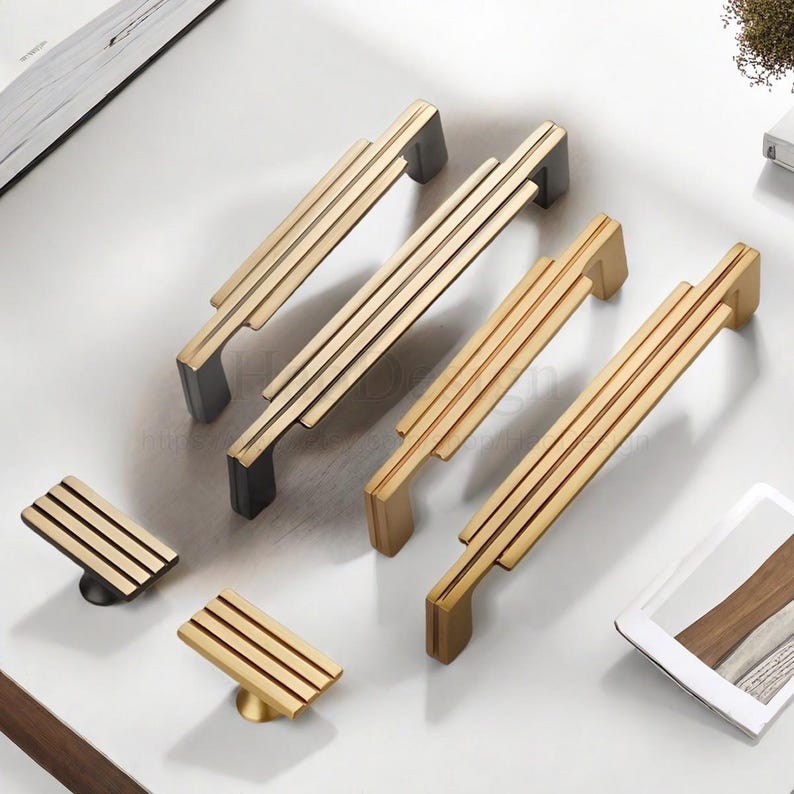 Op de afbeelding: Een verzameling gouden en zwarte kastgrepen en knoppen. De grepen hebben een rechthoekig ontwerp met drie parallelle lijnen, terwijl de knoppen vierkant zijn. De hardware is geplaatst op een wit oppervlak, wat een moderne stijl laat zien.