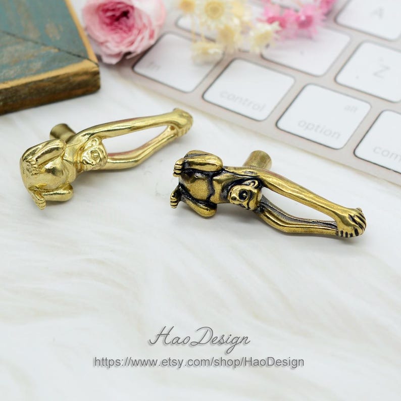 K&ouml;nnte beinhalten: Zwei goldene, affenf&ouml;rmige Schrankkn&ouml;pfe mit detailliertem Design. Der Affe h&auml;lt sich an seinen eigenen Beinen fest und bildet so eine Schlaufe f&uuml;r den Knopf.