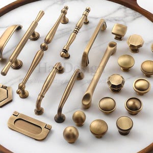 May include: A selection of antique brass cabinet handles and knobs displayed on a white marble surface. The hardware includes a variety of designs, such as simple round knobs and decorative handles, all finished in a warm, aged brass colour.