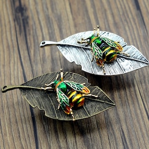 Peut inclure: Deux broches décoratives en forme d'insectes. L'une présente une feuille argentée avec un insecte vert, jaune et orange. L'autre a une feuille de bronze avec un motif d'insecte similaire. Les deux broches sont posées sur un fond en bois.