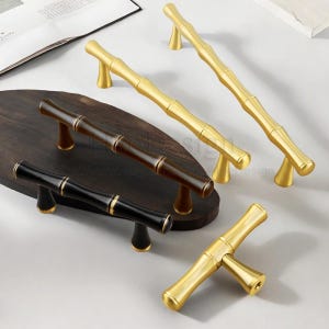 May include: A collection of bamboo-style cabinet pulls in gold, black, and brown. The pulls are arranged on a dark wooden surface and a white surface. The gold pulls have a shiny finish, while the black and brown pulls have a matte finish.
