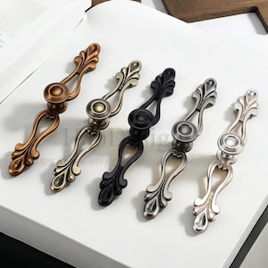 May include: An assortment of decorative cabinet pulls in various metallic finishes. The pulls feature an ornate, symmetrical design with a central knob and curved, stylized handles. Colors include bronze, silver, and black.
