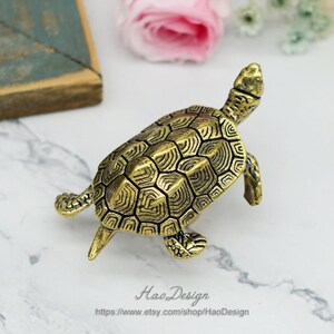Peut inclure: Une figurine de tortue dor&eacute;e avec un motif de carapace d&eacute;taill&eacute;. La tortue est debout sur une surface blanche.