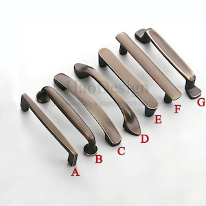 May include: A selection of seven antique bronze cabinet pulls in different styles. The pulls are arranged at an angle, displaying their diverse shapes and finishes. Each pull is labelled with a letter from A to G.