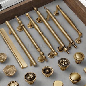 Può includere: Un assortimento di ferramenta per mobili in ottone antico, tra cui varie maniglie e pomelli. Le maniglie variano in lunghezza e design, alcune con dettagli ornati. I pomelli presentano diverse forme e motivi, il tutto in una calda tonalità dorata.