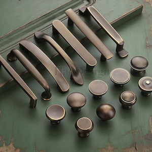 以下が含まれることがあります： アンティーク真鍮製のキャビネット金具の品揃え。取っ手とノブが含まれています。取っ手は様々な形状とサイズがあり、暖かく、使い古された仕上がりです。ノブは丸くドーム型で、クラシックな美しさです。緑色の表面に展示されています。