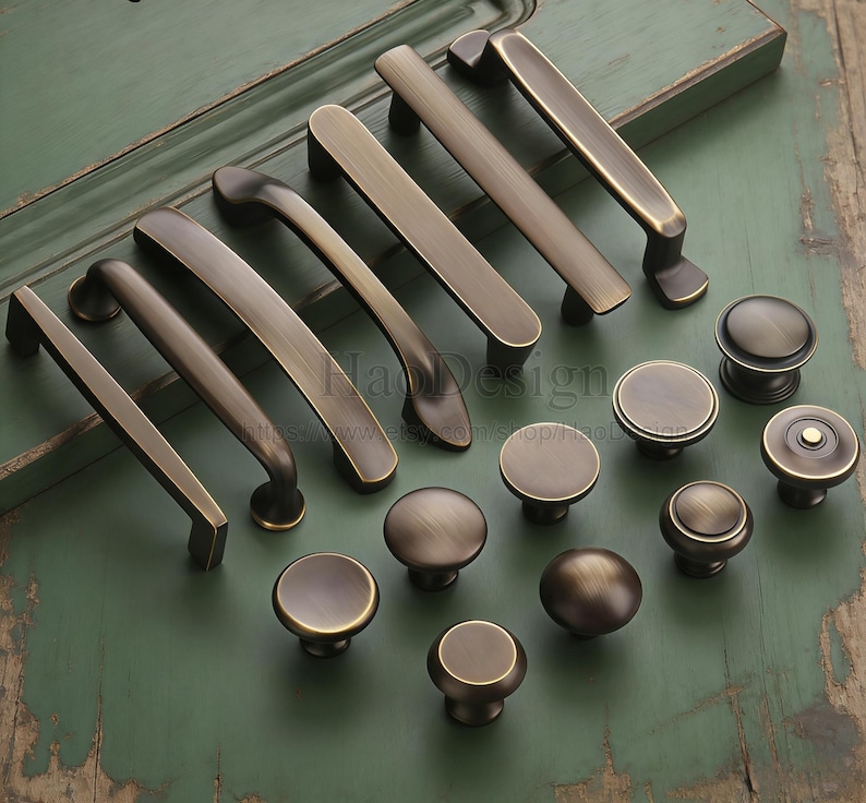May include: A selection of antique brass cabinet handles and knobs. The handles are in various shapes and sizes, with a warm, aged finish. The knobs are round and dome-shaped, providing a classic look. The hardware is displayed on a green surface.