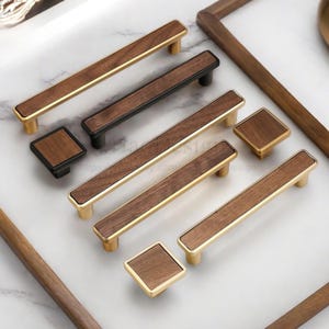 以下が含まれることがあります： 長方形と正方形のプルとノブを備えたキャビネットハードウェアのコレクション。 プルは、木目調の中心部と金または黒の金属トリムを備えています。 ノブは正方形で、木目調の中心部と金または黒の金属トリムが付いています。 白い表面に配置されたハードウェア。