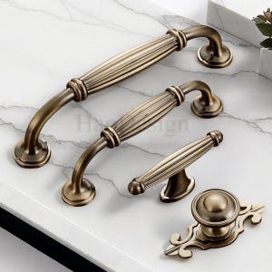 Peut inclure: Ensemble de quincaillerie de meubles en bronze antique. L'ensemble comprend trois tailles de poignées différentes et un bouton décoratif avec une plaque arrière. Les poignées ont un design nervuré et des extrémités incurvées. Le matériel est présenté sur une surface en marbre blanc.